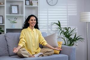 Young beautiful woman meditating at home sitting on sofa in living room in lotus pose, latin american joyful smiling, dreaming and visualizing future achievement results and plans. photo