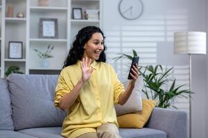 A young curly Indian woman sits on a sofa in a bright living room, holds a phone in her hand, speaks on a call with relatives and friends, greets, smiles and waves her hand at the screen. photo