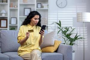 Cheated and sad woman sitting on the sofa inside the house in the living room, Hispanic received a rejection of the transfer, holds a bank credit card in her hands, uses an application on her phone. photo