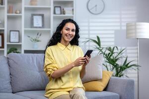 Portrait of a young beautiful hispanic woman at home, smiling and looking at the camera, holding a phone in her hands, using an application on a smartphone, browsing the Internet, typing a message. photo