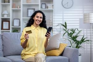 Young beautiful woman sitting on sofa in the living room at home, hisanic girl smiling contentedly using app on phone, using a bank credit card for online shopping, looking at the camera and smiling. photo