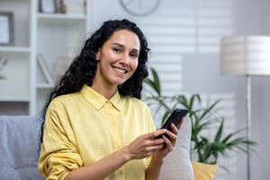 Portrait of a young beautiful hispanic woman at home, smiling and looking at the camera, holding a phone in her hands, using an application on a smartphone, browsing the Internet, typing a message. photo