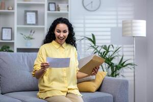 Joyful and happy woman at home received mail envelope with notification, hispanic woman reading good news on paper and happy sitting on sofa in living room . photo