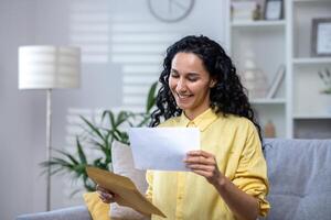 Joyful and happy woman at home received mail envelope with notification, hispanic woman reading good news on paper and happy sitting on sofa in living room . photo