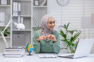 A cheerful Muslim woman wearing a hijab engaged in online teaching, using a laptop and pointing at a small globe, surrounded by books in a well-organized office. photo