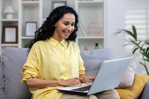 contento Hispano mujer sentado en sofá en brillante vivo habitación con ordenador portátil en regazo, llamar, distancia aprendiendo, trabajo reunión desde hogar. distancia trabajo desde hogar, distancia aprendiendo. foto