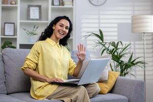 Happy hispanic woman sitting on sofa in bright living room with laptop on lap, studying working student. He waves his hand, smiles, looks at the camera. photo