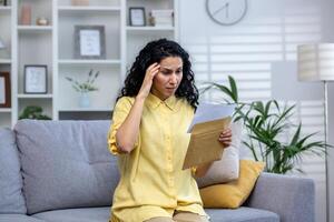 Upset and sad woman at home receiving bad news notification letter, hispanic woman sitting on sofa in living room with mail envelope nervously reading. photo