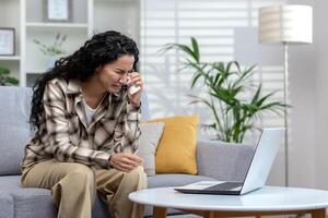 Online consultation with a psychologist, woman crying at home complaining to doctor on call, Hispanic woman with curly hair talking to support service using laptop sitting on sofa at home. photo