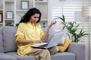 Curly female Hindu student sitting at home on sofa with laptop on lap, unhappy with non-working computer, poor internet connection. Attractive woman having problems working from home. photo