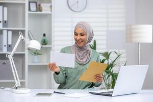 A smiling businesswoman in a hijab is overjoyed as she reads good news from a postal envelope, seated at her modern office desk. photo