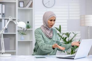 A Muslim woman in a hijab appears stressed and confused while sitting at her desk, dealing with an apparent issue on her laptop in a modern office setting. photo