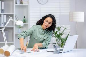 un profesional Hispano mujer en un moderno oficina ajuste meticulosamente trabajos en arquitectónico dibujos junto a su computadora portátil, mostrando atención y pericia. foto