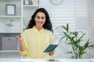 A smiling Hispanic woman conducting an online tutorial with a student, using gestures and holding a notebook in a bright office setting. photo