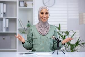 atractivo joven musulmán mujer Hablando enérgicamente en un podcast, direccionamiento el cámara en un bien iluminado, elegante oficina ajuste. foto