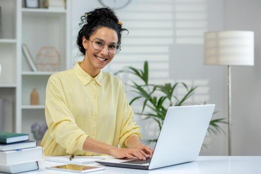 un alegre Hispano mujer vistiendo lentes trabajos en un ordenador portátil en un bien iluminado habitación, rodeado por libros y un planta, emanando productividad y facilidad. foto