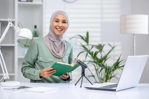 Confident Muslim woman in hijab, recording an audiobook or podcast while holding a green book in a well-lit modern office with plants and a laptop. photo