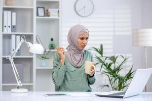 A Muslim businesswoman dealing with a cold at her desk, sipping a soothing hot drink and wiping her runny nose in a modern office setting. photo