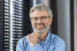 A confident elderly man with grey hair wearing glasses and a blue shirt is smiling in a modern office photo