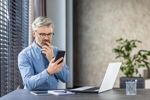 Serious thinking mature gray haired man at workplace inside office, businessman holding phone, thoughtfully reading message browsing email and social media. photo