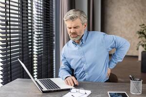 A middleaged man in an office setting experiences discomfort and back pain while working on a laptop photo