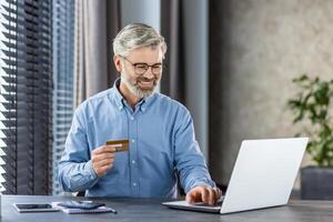 Joyful mature senior businessman at workplace inside office near window with blinds smiling contentedly holding bank credit card in hands, gray haired man using laptop for online shopping. photo