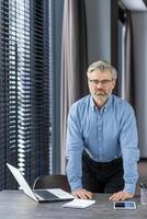 Portrait of a senior businessman standing behind a desk with a laptop, confidently and seriously looking at the camera. photo