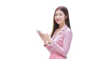 joven asiático profesional trabajando mujer en rosado vestir traje sostiene y mira a su teléfono inteligente felizmente mientras aislado blanco antecedentes. foto