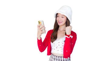 hermosa asiático mujer sonriente con confianza usa rojo Saco y blanco sombrero como santy niña sostiene teléfono inteligente en su mano alegre mientras aislado blanco antecedentes. foto