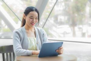 Asian professional business female in light gray long sleeve shirt is looking at tablet or notepad confidently in her hands while she is sitting working at coffee shop among business background city. photo