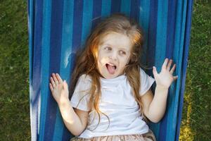 portrait of a little cute girl with long hair resting on a hammock in the garden and showing tongue photo