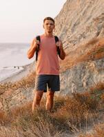 Hiker man in a T-shirt and shorts staying on the ocean coastline at warm sunset. photo