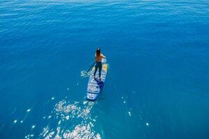 May 20, 2022. Dalaman, Turkey. Sporty woman on stand up paddle board in blue sea. Surfer girl walking on Gladiator SUP board in sea. Aerial view photo