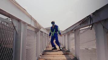 trabajador en un azul uniforme y difícil sombrero caminando en un industrial metal pasarela con barandillas debajo un claro cielo. video