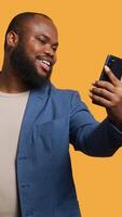 Vertical African american narcissistic man using cellphone to take selfies from all angles. Vain social media user taking photos using phone selfie camera, smiling happily, studio background, camera B video