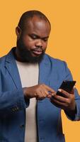 vertical retrato de jubiloso hombre participación Teléfono móvil celebrando, lanzamiento puños en aire y gritando. Encantado africano americano persona leyendo bueno Noticias en móvil teléfono, aplausos, estudio fondo, cámara un video