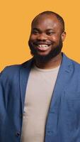 Vertical African american man cheerfully doing salutation hand gesture. Portrait of happy BIPOC person raising arm to greet someone, gesturing, isolated over yellow studio background, camera A video