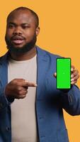 Vertical Smiling african american man presenting cellphone with green screen display, isolated over studio background. Cheerful BIPOC person creating promotion with blank copy space mockup phone, camera A video