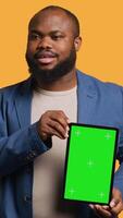 Vertical Amused african american man laughing while presenting tablet with green screen display, studio background. Joyous BIPOC person has fun during promotion for blank copy space mockup device, camera A video