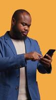 Vertical Portrait of joyous man holding cellphone celebrating, throwing fists in air and screaming. Delighted african american person reading good news on mobile phone, cheering, studio background, camera B video