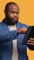 Vertical Man happily scrolling on tablet touchscreen to check social media feed. African american person browsing internet websites, entertained by content, studio background, camera A video