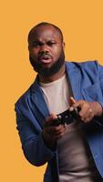 verticaal portret van vrolijk bipoc Mens spelen spellen Holding controleur, studio achtergrond. gamer lachend, hebben pret door deelnemen in pvp online multiplayer spel gebruik makend van troosten systeem, camera b video