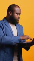 Vertical African american man using laptop to do business related tasks for company, isolated over studio background. Office clerk doing budgeting, forecasting, and financial analysis on notebook, camera B video