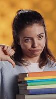 Vertical Portrait of woman with pouting expression holding pile of books, showing disproval of reading hobby. Sulky lady with stack of novels doing thumbs down hand gesturing, studio background, camera A video