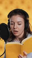 Vertical Woman does voiceover reading of book to produce audiobook using dramatic acting. Narrator uses storytelling skills and expressive sounds while producing recording of novel, studio background, camera A video