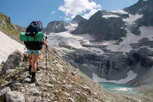 turistas en pantalones cortos con enorme mochilas con mochilero almohadillas y trekking polos caminando arriba morena de montaña. en antecedentes blanco picos y cascada fluir dentro un azur montaña lago en soleado día foto