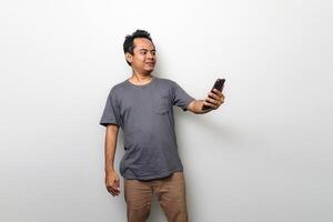 un asiático hombre en gris es sorprendido cuando él mira a un teléfono inteligente aislado en blanco antecedentes foto