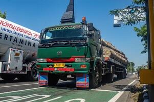 un remolque camión que lleva grande registros desde el bosque o un Inicio sesión camión paso en un urbano carretera, Indonesia, dieciséis mayo 2024. foto