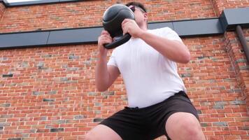 Sport und Freizeit. stark kaukasisch Mann Arbeiten aus mit Kettlebell im das Park, Backstein Mauer Hintergrund video