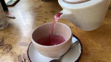 Hibiscus red tea is poured into a glass cup. Pour freshly brewed tea from a teapot into clear double-walled cup. The process of pouring hot fruit red tea with hibiscus. Hibiscus tea on wooden table video